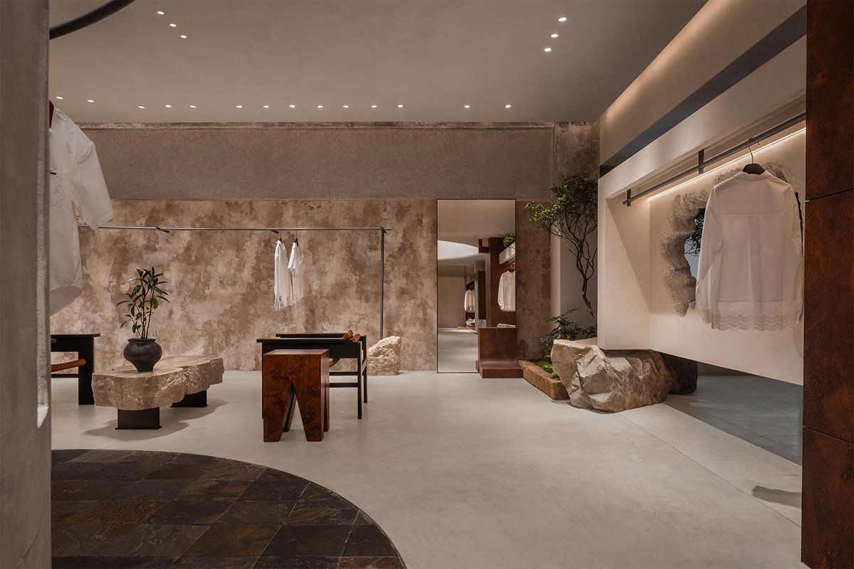 Display area in the Hé showroom with display tables, textured wall, full-height mirror, natural stones and suspended garments